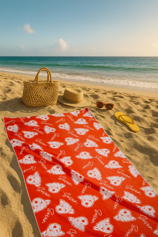 Red beach towel with white patterns on a sandy beach with a straw bag, hat, and sandals.