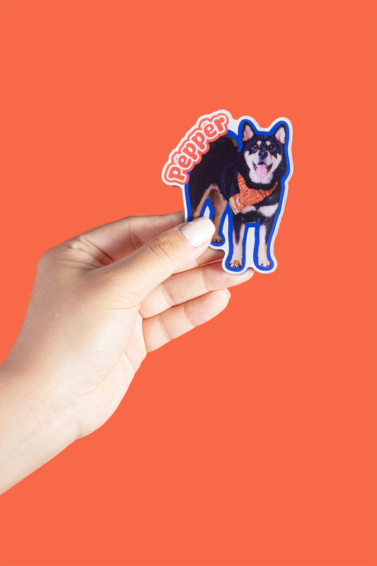 Hand holding a sticker of a dog with 'Pepper' text against an orange background