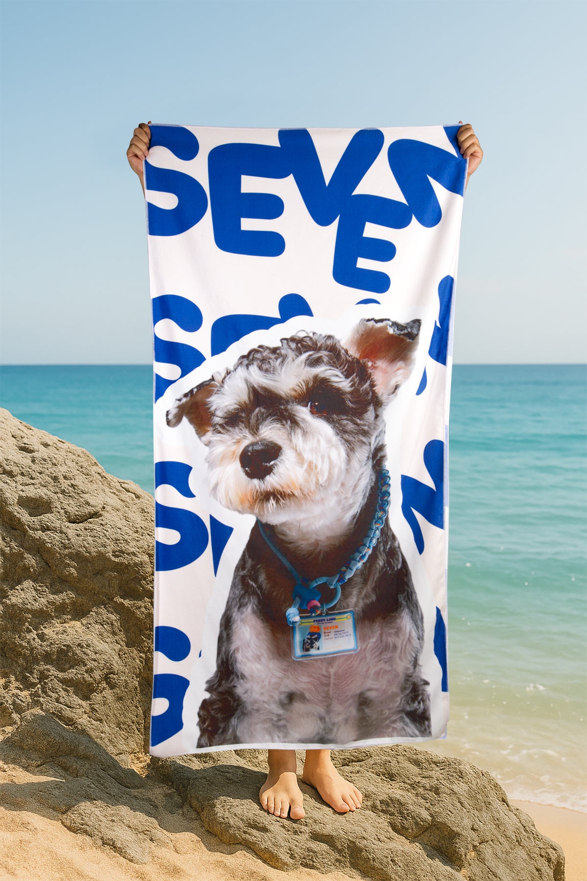 Person holding a beach towel with a dog's face and 'SEVEN' printed on it at the beach.