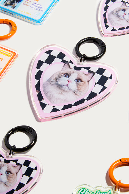 Heart-shaped keychain with a checkered border featuring a cat's face, surrounded by other keychains on a light background.