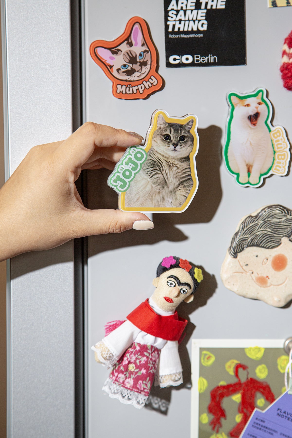 Hand placing a cat-shaped magnet on a refrigerator with other magnets and a doll attached.