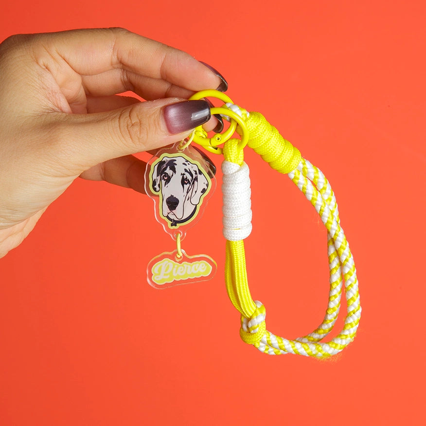 A hand holds a bright yellow and white braided rope dog leash with a yellow ring, white wrapped section, and two charms: a black and white dog face and a clear 'Pierce' nameplate, set against a solid orange background.